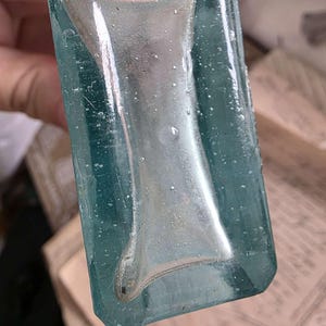 May include: A vintage, rectangular glass bottle with a teal tint. The bottle features a silver-colored, hourglass-shaped design in the center. The glass has a slightly textured surface with small bubbles, suggesting an antique or handcrafted origin.