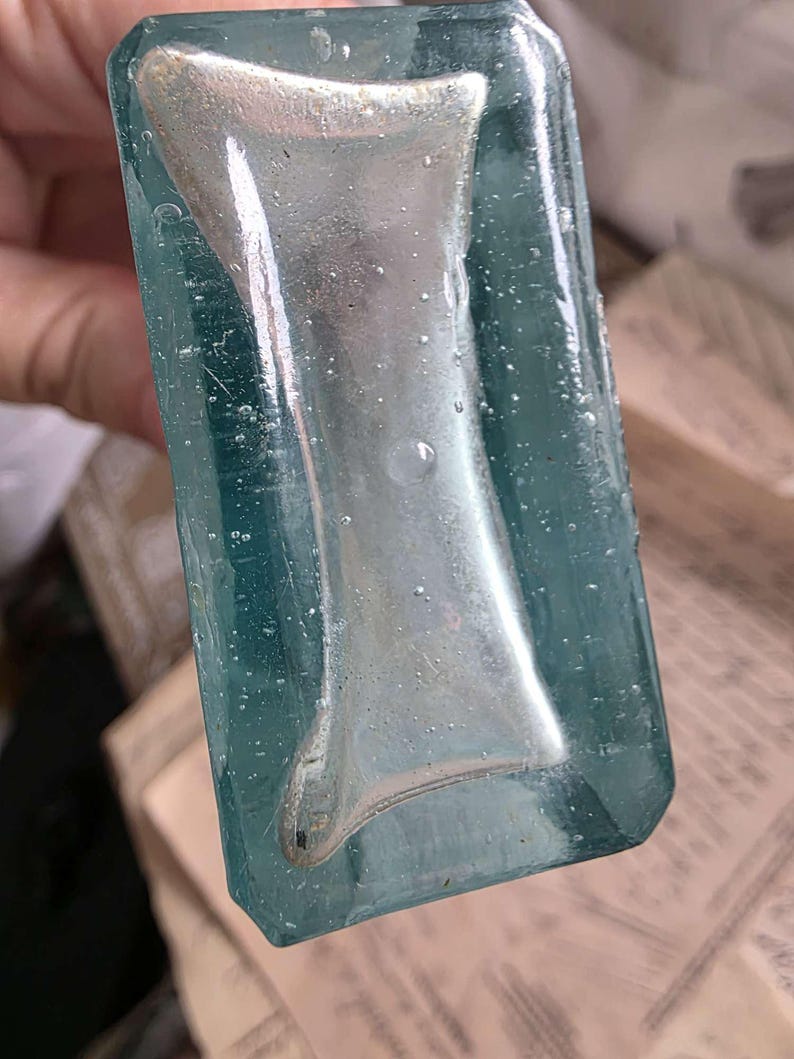 May include: A vintage, rectangular glass bottle with a teal tint. The bottle features a silver-colored, hourglass-shaped design in the center. The glass has a slightly textured surface with small bubbles, suggesting an antique or handcrafted origin.