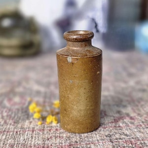 May include: A small, cylindrical, antique brown stoneware bottle with a narrow neck and a rounded rim. The bottle has a textured surface and is slightly worn, suggesting age. A few yellow flowers are in the foreground.
