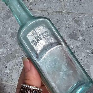 May include: An antique, rectangular, light blue glass bottle with the embossed word "DAVIS". The bottle has a long neck and a slightly iridescent finish. The bottle is held by a person wearing a ring with oval-shaped stones.