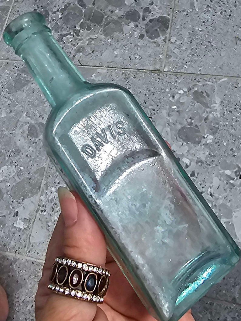 May include: An antique, rectangular, light blue glass bottle with the embossed word "DAVIS". The bottle has a long neck and a slightly iridescent finish. The bottle is held by a person wearing a ring with oval-shaped stones.