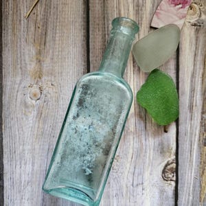 May include: An antique, rectangular, light blue glass bottle with a textured surface. Beside the bottle are two pieces of sea glass, one green and one gray, and a small piece of paper with a floral design. The items are arranged on a wooden surface.