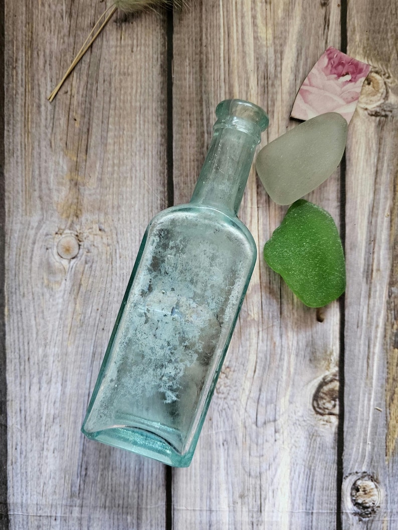 May include: An antique, rectangular, light blue glass bottle with a textured surface. Beside the bottle are two pieces of sea glass, one green and one gray, and a small piece of paper with a floral design. The items are arranged on a wooden surface.