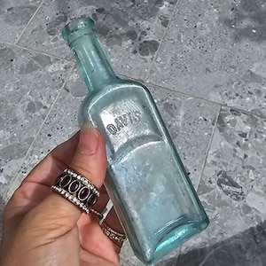 May include: A vintage, rectangular, light blue glass bottle with the embossed word "DAVIS". The bottle has a narrow neck and a slightly iridescent finish. The bottle is held in a hand with multiple rings.