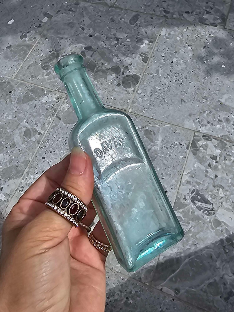 May include: A vintage, rectangular, light blue glass bottle with the embossed word "DAVIS". The bottle has a narrow neck and a slightly iridescent finish. The bottle is held in a hand with multiple rings.