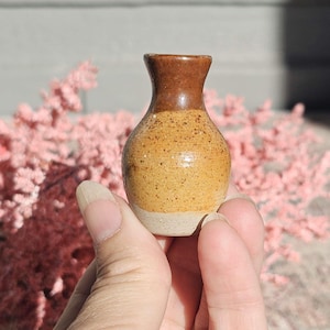 Puede incluir: Un pequeño jarrón de cerámica hecho a mano, con un cuerpo moteado de color beige y un borde marrón oscuro. La base es de un color más claro, sin esmaltar. El jarrón se sostiene en la mano de una persona, mostrando su tamaño y textura.