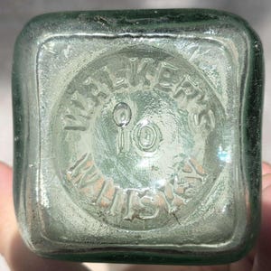May include: Close-up of the bottom of a vintage, square-shaped, light green glass bottle. The embossed text reads "WALKER'S" in a circular pattern, with "10" in the center and "WHISKY" below. The glass has a slightly textured appearance.