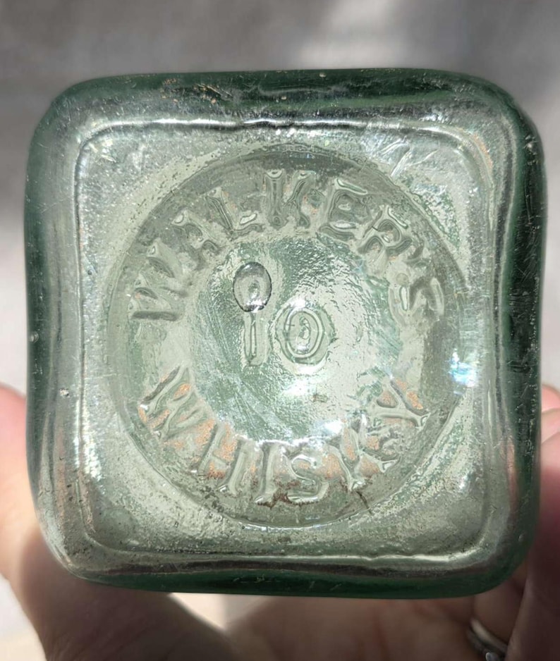 May include: Close-up of the bottom of a vintage, square-shaped, light green glass bottle. The embossed text reads "WALKER'S" in a circular pattern, with "10" in the center and "WHISKY" below. The glass has a slightly textured appearance.