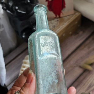 May include: An antique, rectangular, light blue glass bottle with the embossed word "DAVIS". The bottle has a narrow neck and a slightly faded appearance. The bottle is held in a hand with a decorative ring.