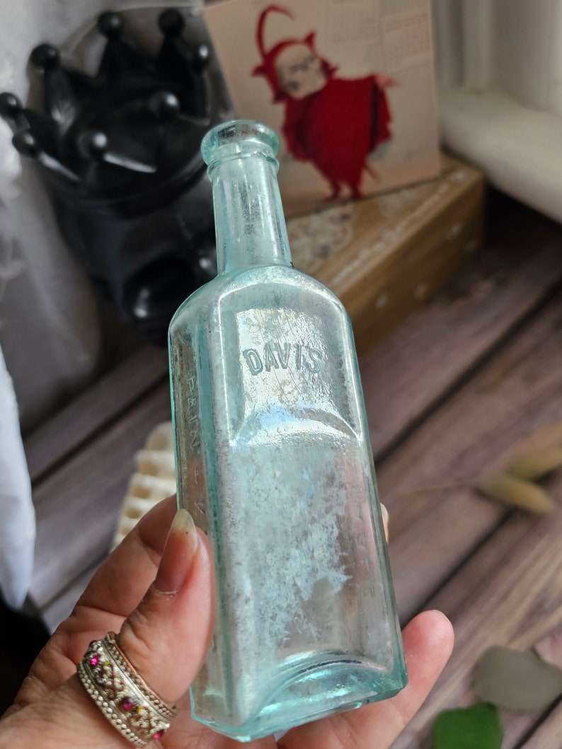 May include: An antique, rectangular, light blue glass bottle with the embossed word "DAVIS". The bottle has a narrow neck and a slightly faded appearance. The bottle is held in a hand with a decorative ring.