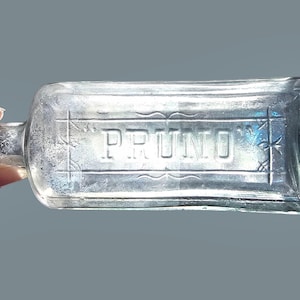 Może przedstawiać: Przezroczysta szklana butelka z wytłoczonym napisem "PRUNO". Prostokątna butelka ma zaokrągloną szyjkę i płaską podstawę. Butelka jest trzymana przez dłoń ze srebrnym pierścionkiem.
