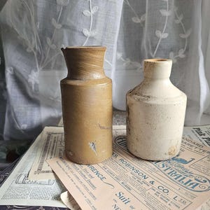 Puede incluir: Dos botellas de cerámica antiguas en tonos beige y blanquecino sobre un periódico vintage. La botella más alta es cilíndrica con la parte superior ligeramente ensanchada. La botella más corta tiene un cuerpo redondeado y un cuello estrecho.