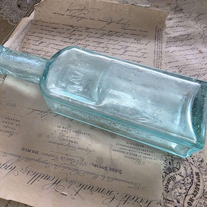 May include: A vintage, rectangular, light blue glass bottle with a long neck, resting on aged paper with French text. The bottle has a slightly faded appearance, suggesting age and use.