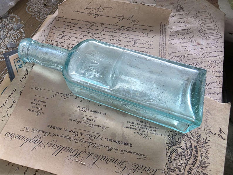 May include: A vintage, rectangular, light blue glass bottle with a long neck, resting on aged paper with French text. The bottle has a slightly faded appearance, suggesting age and use.