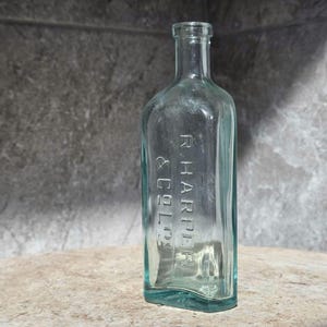 May include: A clear, antique glass bottle with a rectangular shape and a narrow neck. The bottle has the embossed text "R. HARPER & CO." on the front. The glass has a slight teal tint. The bottle is displayed on a light brown surface.