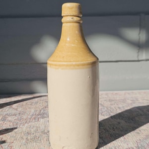 Könnte beinhalten: Eine Vintage-Steinzeugflasche mit zylindrischem Körper und abgerundeter Schulter. Die Flasche hat einen hellbeigen Sockel und einen hellbraunen Hals und Verschluss. Sie wird wahrscheinlich zur Aufbewahrung von Flüssigkeiten verwendet, ca. 25 cm hoch.