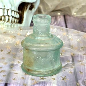Puede incluir: Una pequeña botella de vidrio verde claro de estilo antiguo, con base redondeada y cuello cilíndrico. La botella tiene un aspecto ligeramente desgastado, lo que sugiere antigüedad. El fondo incluye una calavera y una tela con patrones de estrellas doradas.