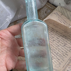 May include: A vintage, light blue glass bottle with a rectangular body and a long neck. The bottle is held in a hand, and the background includes antique papers and a white lace curtain. The bottle's color is a pale aqua.