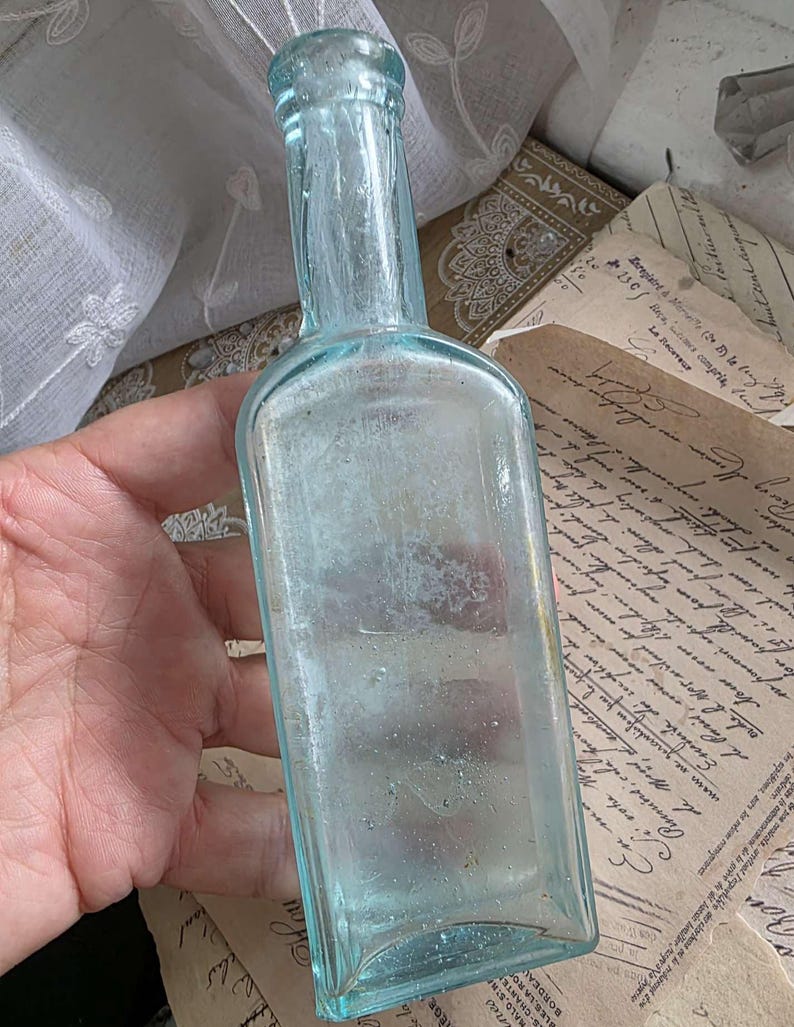 May include: A vintage, light blue glass bottle with a rectangular body and a long neck. The bottle is held in a hand, and the background includes antique papers and a white lace curtain. The bottle's color is a pale aqua.