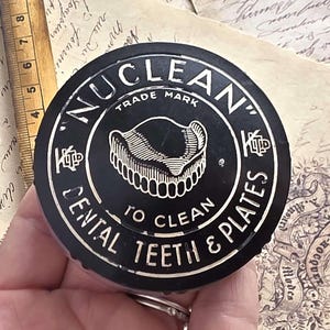 May include: Black round container with white text reading "NUCLEAN" and "DENTAL TEETH & PLATES". The container has an image of dentures. The words "TRADE MARK" are also visible.