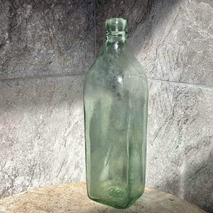 May include: A clear, antique-style glass bottle with a square base and a narrow neck. The bottle has a slight green tint and is displayed against a textured gray background. The bottle's design suggests it may have been used for storing liquids.