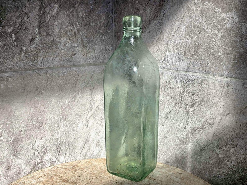 May include: A clear, antique-style glass bottle with a square base and a narrow neck. The bottle has a slight green tint and is displayed against a textured gray background. The bottle's design suggests it may have been used for storing liquids.