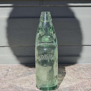 Puede incluir: Una botella de vidrio transparente vintage con un ligero tinte verde. La botella tiene una forma única con un cuello estrecho y un cuerpo redondeado. El texto "BOXHALL WINDSOR" está grabado en la botella. Probablemente una botella antigua de refresco o agua mineral.