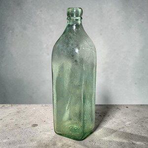 May include: A vintage, rectangular-shaped, light green glass bottle. The bottle has a rounded neck and a flat base. The glass has a slightly textured appearance, suggesting it is an antique or a replica of an antique bottle.