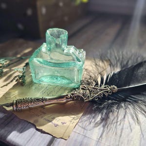 Könnte beinhalten: Ein antikes Glas-Tintenfass mit grüner Tönung, eine schwarze Federkiel mit einem verzierten silbernen Halter und ein Stück Papier mit handschriftlichem Text.