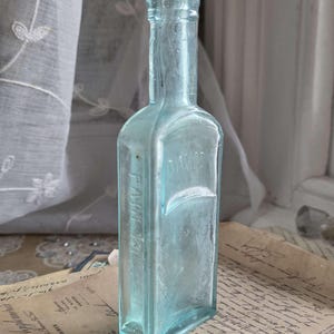 May include: A vintage, rectangular, light blue glass bottle with a long neck. The bottle has the embossed text "Farm Ville" on the side. The bottle is displayed on a stack of aged papers, with a white curtain in the background.