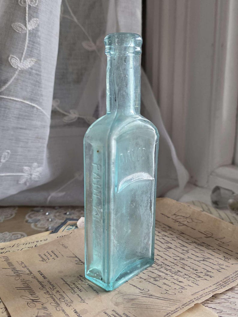 May include: A vintage, rectangular, light blue glass bottle with a long neck. The bottle has the embossed text "Farm Ville" on the side. The bottle is displayed on a stack of aged papers, with a white curtain in the background.