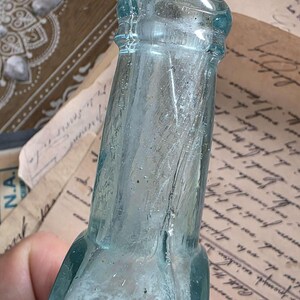 May include: A vintage, light blue glass bottle with a textured surface. The bottle has a narrow neck and a rounded base. The glass has a slight tint and appears to be antique. The bottle is held against a background of aged paper.