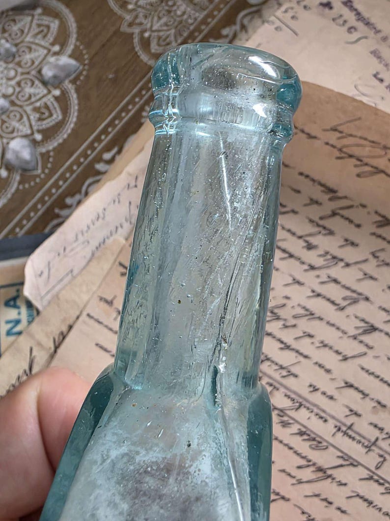 May include: A vintage, light blue glass bottle with a textured surface. The bottle has a narrow neck and a rounded base. The glass has a slight tint and appears to be antique. The bottle is held against a background of aged paper.