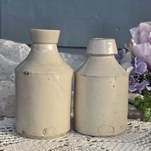 May include: Two antique, off-white stoneware bottles with a cylindrical body and a short neck. The bottles have a slightly aged appearance, with subtle variations in color and texture. The base of each bottle features a small, embossed design.