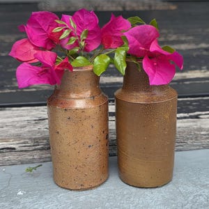 Puede incluir: Dos jarrones de cerámica marrón con flores de buganvillas de color rosa brillante y hojas verdes. Los jarrones tienen una textura rústica y moteada y una forma cilíndrica. Las flores añaden un contraste vibrante a los tonos terrosos de los jarrones.