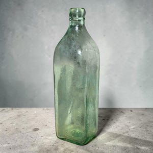 May include: A vintage, rectangular-shaped, light green glass bottle. The bottle has a rounded neck and a flat base. The glass has a slightly textured appearance, suggesting it is an antique or a replica of an antique bottle.