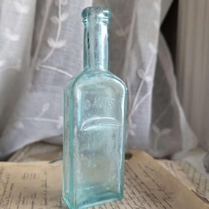 May include: A vintage, rectangular, light blue glass bottle with the embossed word "DAVIS". The bottle has a long neck and a slightly rounded top. It is displayed on a vintage paper with faded text, suggesting an antique apothecary item.