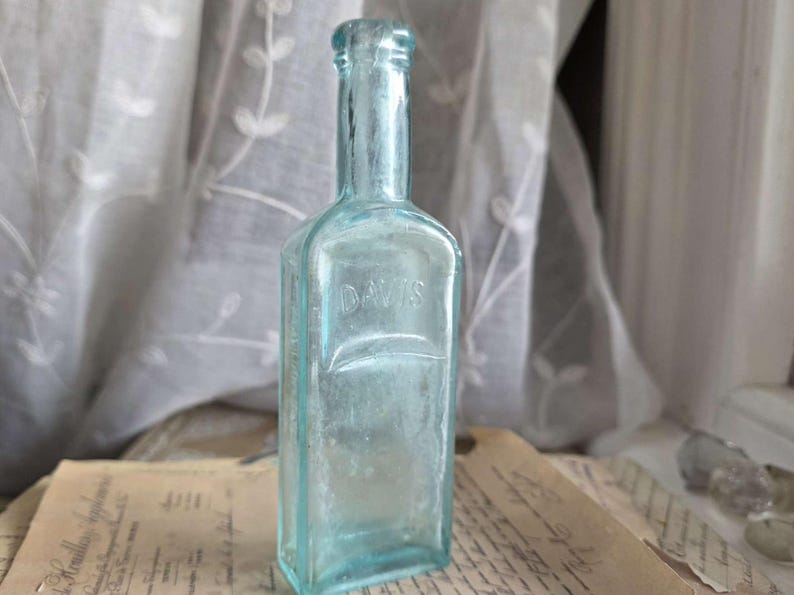 May include: A vintage, rectangular, light blue glass bottle with the embossed word "DAVIS". The bottle has a long neck and a slightly rounded top. It is displayed on a vintage paper with faded text, suggesting an antique apothecary item.