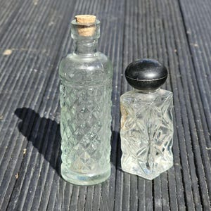 Pode incluir: Duas garrafas de vidro vintage com desenhos em losangos. Uma garrafa tem uma rolha de cortiça e a outra tem uma rolha preta e arredondada. As garrafas são transparentes e estão sobre uma superfície escura e texturizada.