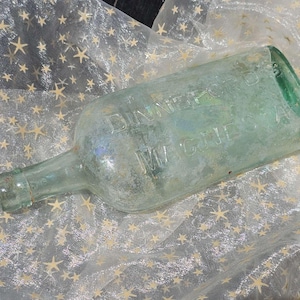 Puede incluir: Se muestra una botella antigua de vidrio transparente con el texto en relieve "Dinner's M. Cure". La botella tiene un cuerpo rectangular y una base redondeada. La botella está sobre una tela blanca con patrones de estrellas doradas.