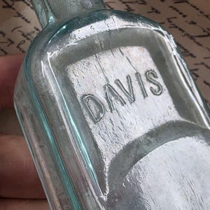 May include: A vintage, light blue glass bottle with the embossed word "DAVIS" on the front. The bottle has a rectangular shape with rounded edges and a long neck. The glass has a slightly textured appearance.