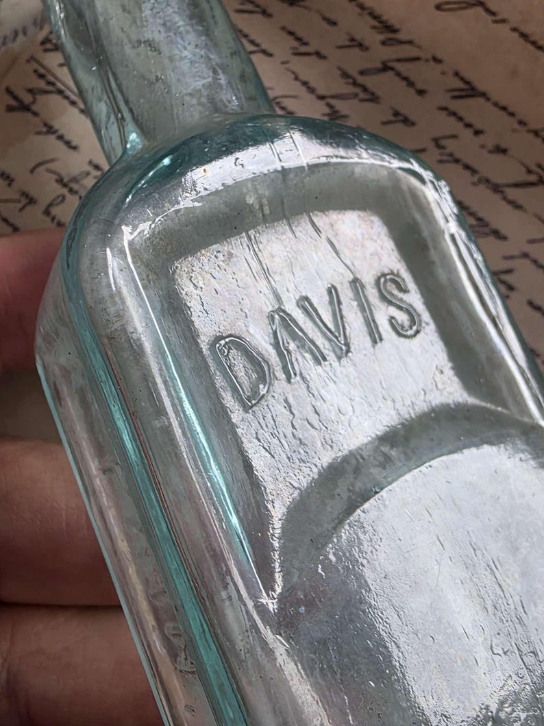 May include: A vintage, light blue glass bottle with the embossed word "DAVIS" on the front. The bottle has a rectangular shape with rounded edges and a long neck. The glass has a slightly textured appearance.