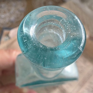 May include: A vintage, light blue glass bottle with a square base and a round, open neck. The bottle has a slightly textured surface and is partially transparent, allowing light to pass through. The bottle's design suggests it may have been used for medicine or perfume.