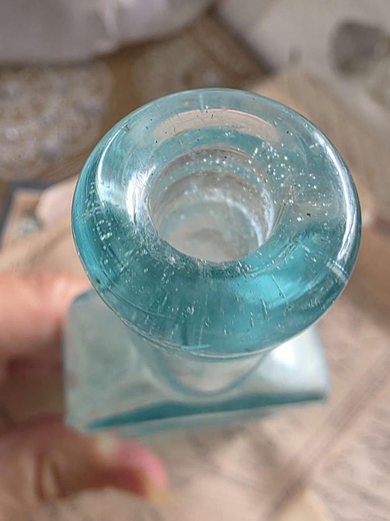 May include: A vintage, light blue glass bottle with a square base and a round, open neck. The bottle has a slightly textured surface and is partially transparent, allowing light to pass through. The bottle's design suggests it may have been used for medicine or perfume.