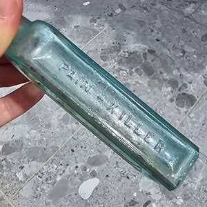 May include: A vintage, rectangular, light blue glass bottle with the embossed text "PAIN KILLER". The bottle has a slightly rounded neck and is held by a hand. The glass has a slight greenish tint.