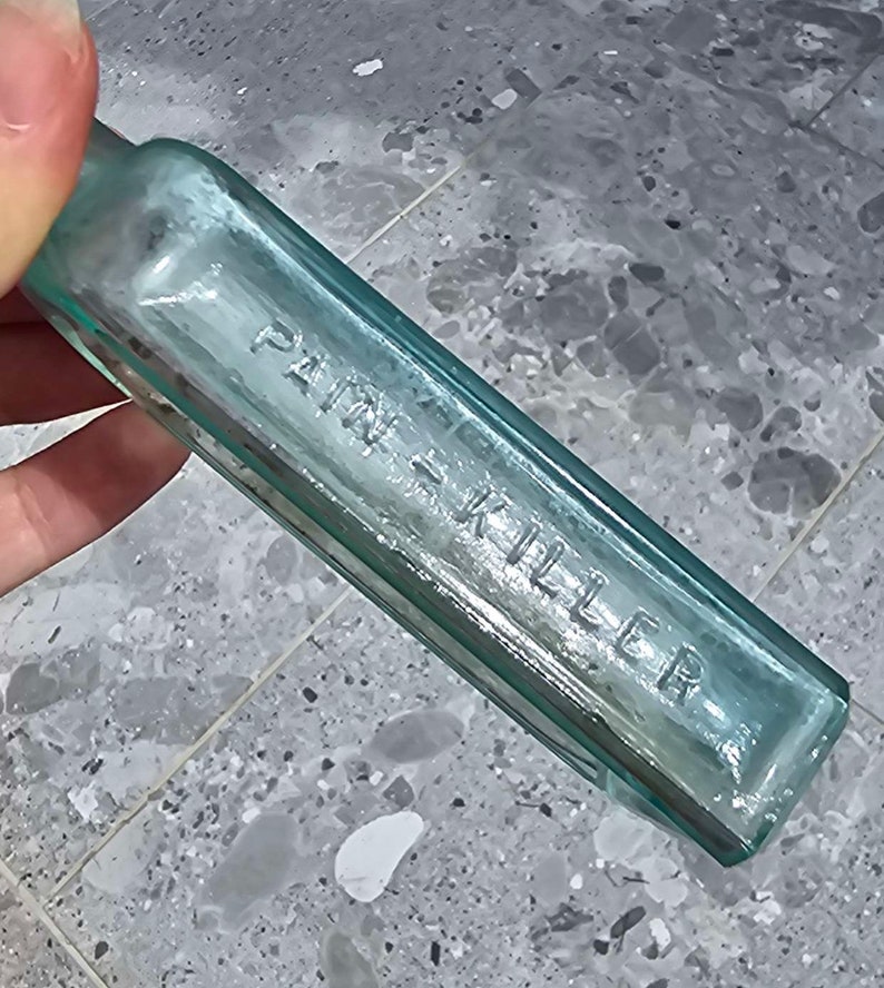 May include: A vintage, rectangular, light blue glass bottle with the embossed text "PAIN KILLER". The bottle has a slightly rounded neck and is held by a hand. The glass has a slight greenish tint.