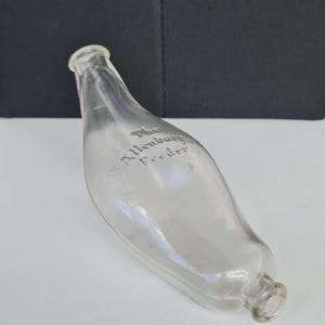 May include: Clear glass bottle with the text "The Allenbury Feeder" etched on the side. The bottle has a curved shape with a narrow neck and a rounded base. The bottle is empty and appears to be vintage.