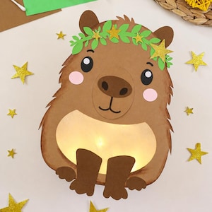 Könnte beinhalten: Ein Capybara-Bastelobjekt aus Papier in Braun mit einer Krone aus grünen Blättern und goldenen Glitzersternen, rosa Wangen und einem leuchtenden Bauch. Das Capybara ist von mehreren kleinen, einzelnen goldenen Glitzersternen umgeben.