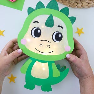 May include: A green paper craft dinosaur character with a glittery head crest and tail. The face and belly sections are translucent, illuminated by internal warm white LED lights. The character features large black eyes, pink cheeks, and a smile.