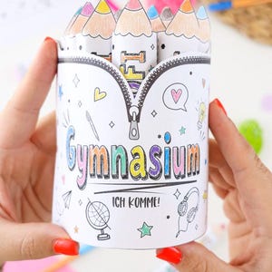 Peut inclure: Un porte-crayons cylindrique en papier blanc avec un motif de fermeture éclair imprimé sur le dessus et des illustrations de crayons de couleur. Le texte Gymnasium et ICH KOMME! est imprimé sur le devant avec des icônes comme des étoiles et des cœurs.
